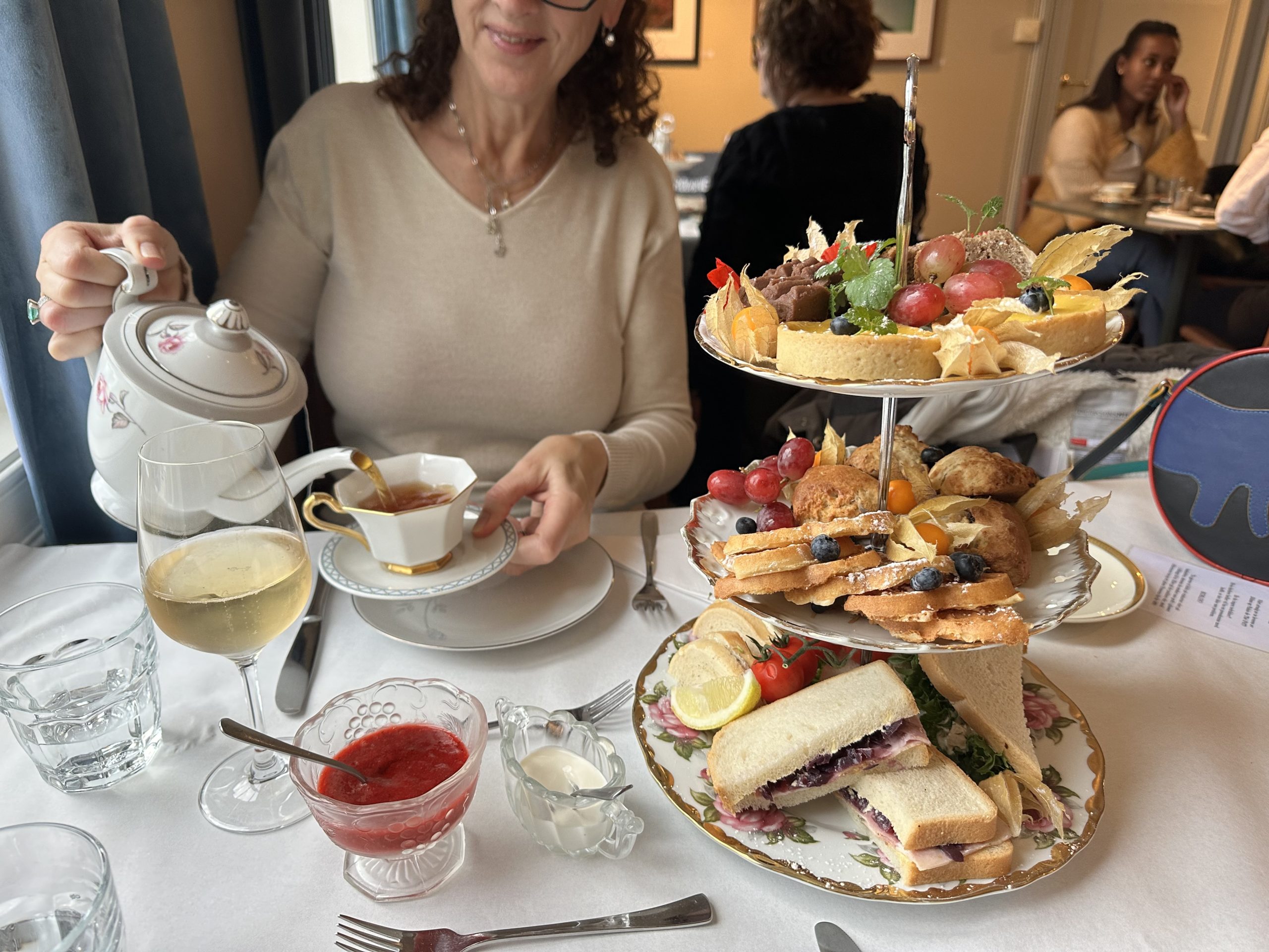 Afternoon tea på Jæren, kvinne heller te I en tekopp på et bord med kaker i et etasjefat.